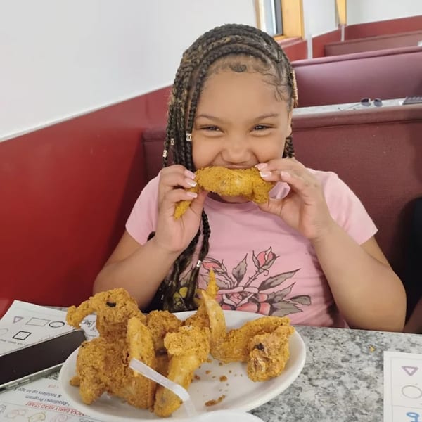 A young girl in a pink shirt happily eats a piece of fried chicken at Broadway FAMILY Grill.
