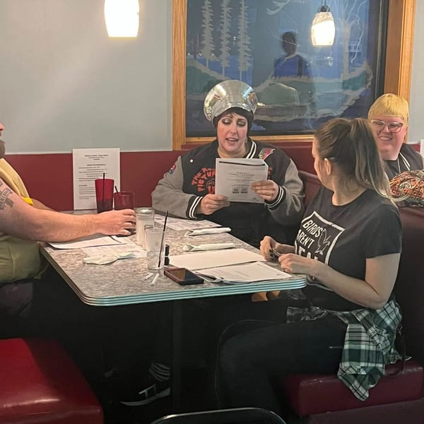 Four people seated at a diner booth, with one person in a shiny hat reading a menu.