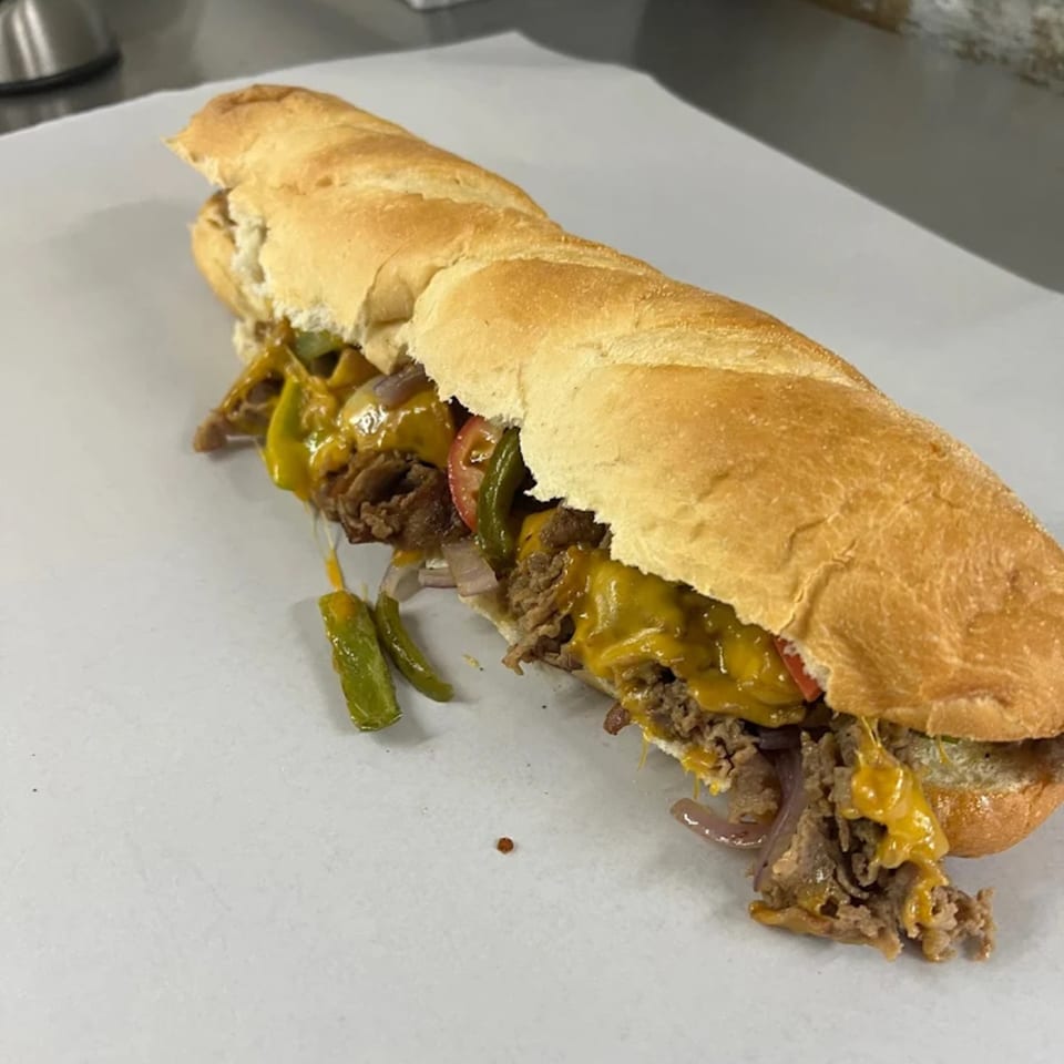 A large, cheesy Philly cheesesteak sandwich with peppers and onions on a white surface.