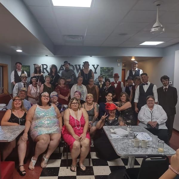 Large group of people in 1920s attire posing in Broadway FAMILY Grill, with checkered floors.