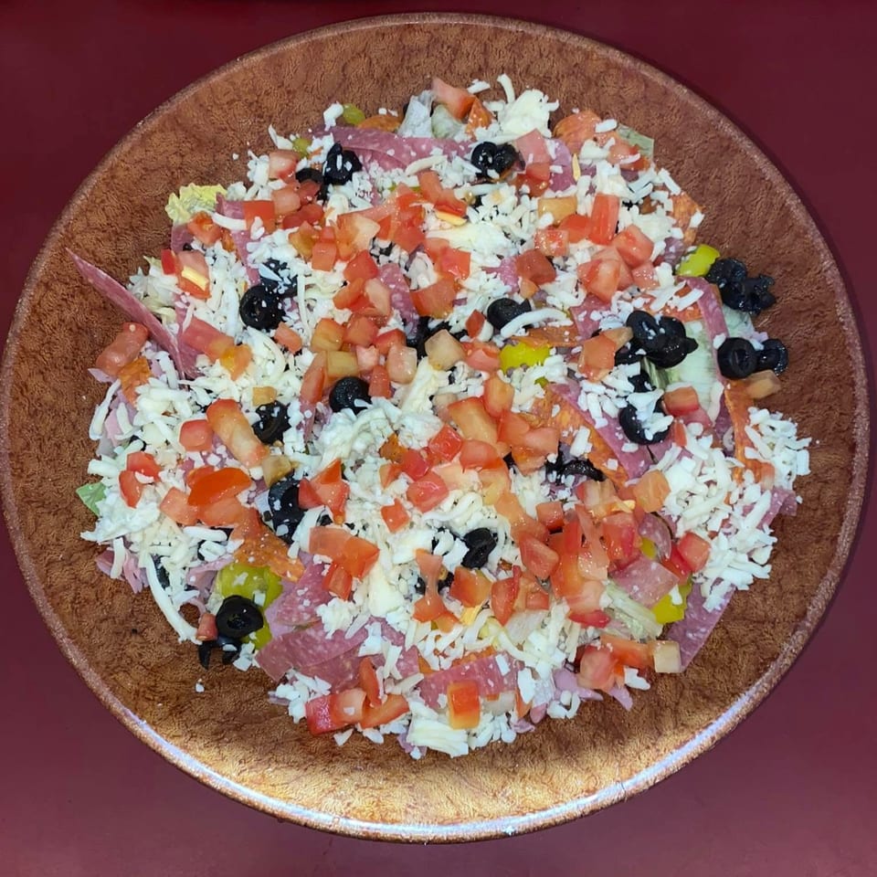 A large salad with shredded cheese, diced tomatoes, black olives, and various meats in a brown bowl.