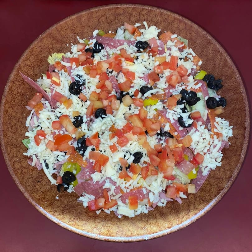 A large salad with shredded cheese, diced tomatoes, black olives, and various meats in a brown bowl.